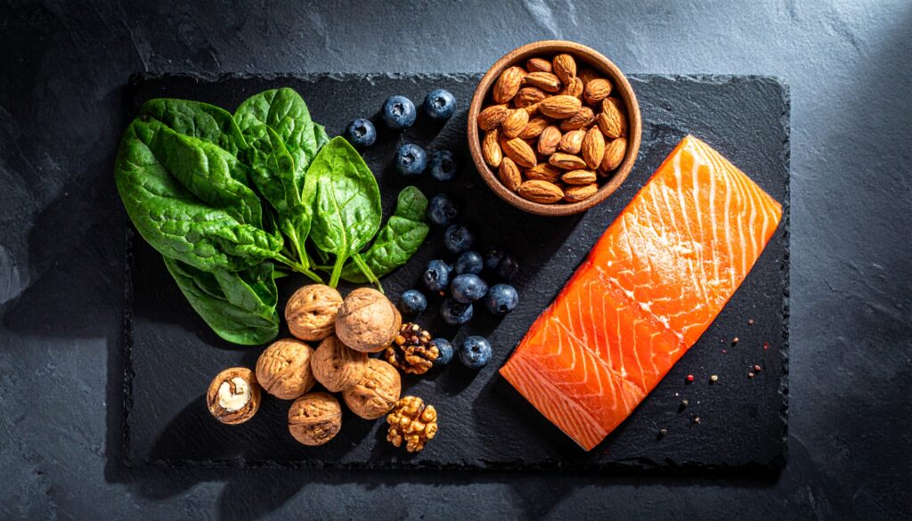 Auf einer dunklen Schieferplatte sind hochwertige, natürliche Lebensmittel wie ein Stück Lachs, eine Handvoll Walnüsse, dunkelgrünes Blattgemüse und einige Blaubeeren minimalistisch und elegant arrangiert. Das Licht kommt von der Seite und hebt die Texturen hervor.