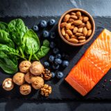Auf einer dunklen Schieferplatte sind hochwertige, natürliche Lebensmittel wie ein Stück Lachs, eine Handvoll Walnüsse, dunkelgrünes Blattgemüse und einige Blaubeeren minimalistisch und elegant arrangiert. Das Licht kommt von der Seite und hebt die Texturen hervor.